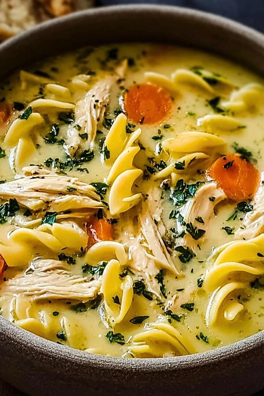A bowl of creamy chicken noodle soup with vegetables and herbs.